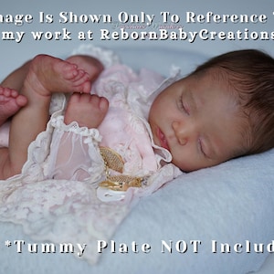 Może przedstawiać: Realistyczna lalka reborn, śpiąca spokojnie na białej poduszce. Lalka ubrana jest w różowy strój z koronkowymi wykończeniami. Obraz zawiera tekst, który mówi "This Image Is Shown Only To Reference The Kit" i "View my work at RebornBabyCreations.com".
