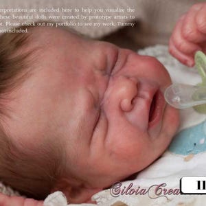 Może przedstawiać: Lalka noworodka z zamkniętymi oczami i smoczkiem w buzi. Lalka ma szczegółowe odcienie skóry i delikatne włosy. Lalka leży na białym i niebieskim wzorzystym kocu. Widoczny jest tekst "112-43".