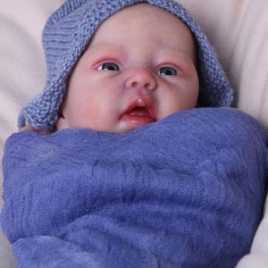 Może przedstawiać: Lalka reborn owinięta w miękki, niebiesko-fioletowy koc. Lalka ma na sobie pasujący dzianinowy czepek. Lalka ma jasną skórę, niebieskie oczy i lekko otwarte usta. Tło jest w miękkim, neutralnym kolorze.