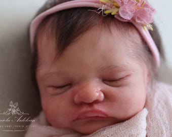 May include: A close-up of a sleeping baby doll with a pink headband and a flower accent. The doll has realistic features and is wearing a white fabric.