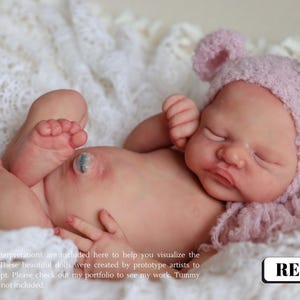 Op de afbeelding: Een levensechte babypop met gesloten ogen, met een roze gebreide muts met berenoren en liggend op een witte kanten deken. De pop heeft realistische huidtinten en kenmerken. De tekst "REF-001" staat rechtsonder.