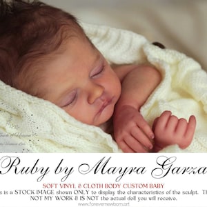 Może przedstawiać: Realistyczna lalka dla dziecka o imieniu "Ruby" autorstwa Mayry Garza, owinięta w kremowy, dziergany kocyk. Lalka ma zamknięte oczy, szczegółowe rysy twarzy i brązowe włosy. Tekst na zdjęciu brzmi "SOFT VINYL & CLOTH BODY CUSTOM BABY".