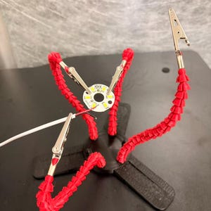 May include: A black base with four flexible red articulated arms, each ending in a metal alligator clip. One clip holds a small circular white circuit board with multiple LED components, while another clip holds a thin white and red wire.