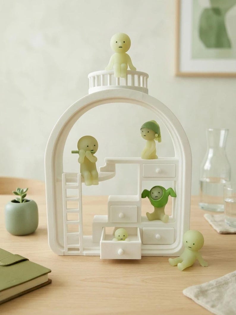 May include: A white decorative shelf unit with a curved top and multiple small drawers, housing several small, light green figurines. One figurine sits atop the unit, while others are positioned on the stairs and in the drawers. A small potted plant is visible on the left.