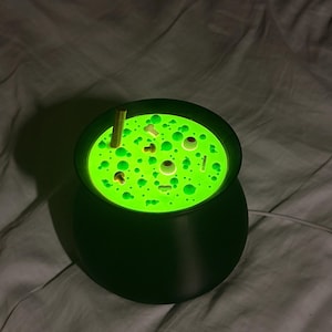 Może przedstawiać: Czarna lampa w kształcie kociołka emitująca z góry jasne, neonowo-zielone światło. Powierzchnia lampy zawiera małe, wypukłe kształty, w tym gałki oczne, kości i grzyby, tworząc efekt bulgoczącego płynu z białym przewodem zasilającym.