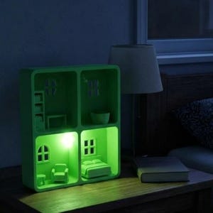 May include: A green, illuminated dollhouse night light with miniature furniture and windows. The night light is on a wooden nightstand next to a lamp and a book. The light emits a soft green glow.