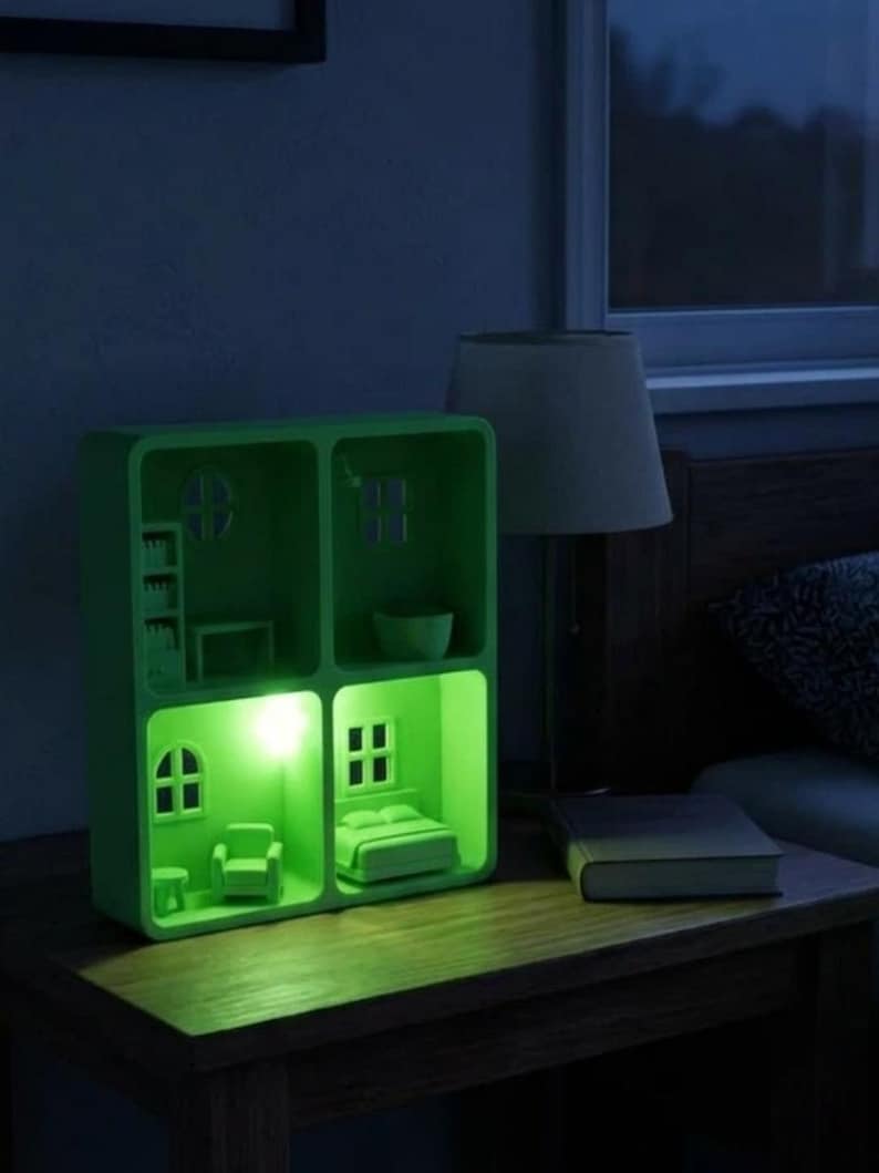 May include: A green, illuminated dollhouse night light with miniature furniture and windows. The night light is on a wooden nightstand next to a lamp and a book. The light emits a soft green glow.