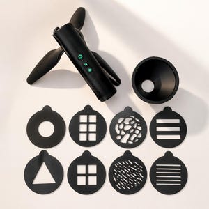 May include: A black handheld projector device with three buttons featuring green icons, a circular lens attachment, and eight circular black discs with various cut-out patterns including a triangle, squares, lines, and organic shapes.