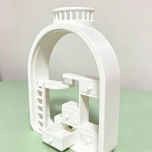 May include: A white miniature dollhouse-style structure with an arched frame. It features a ladder, a desk, and several drawers, one of which is open. The top has a circular design with vertical bars. The overall design is minimalist and modern.