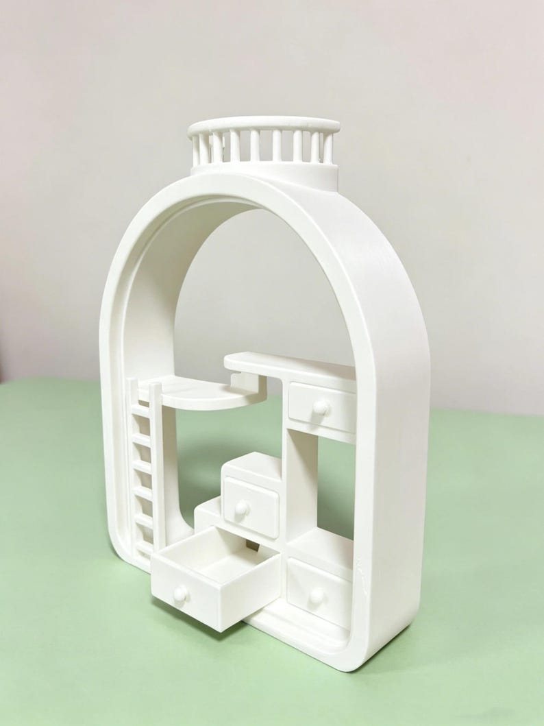 May include: A white miniature dollhouse-style structure with an arched frame. It features a ladder, a desk, and several drawers, one of which is open. The top has a circular design with vertical bars. The overall design is minimalist and modern.