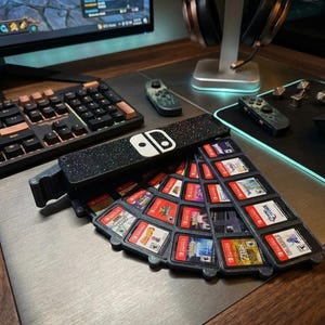 Könnte beinhalten: Ein schwarzer, fächerförmiger Aufbewahrungskoffer für Spielkassetten mit gesprenkelter Oberfläche und dem weißen Nintendo Switch-Logo auf dem Deckel. Der Koffer ist fächerartig geöffnet und zeigt mehrere Fächer mit verschiedenen Spielkassetten.
