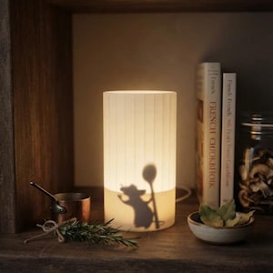 May include: A cylindrical white lamp with vertical ribbed texture, featuring a dark silhouette of a mouse holding a large spoon projected onto its surface. The lamp emits a warm glow, illuminating the surrounding space with soft light.