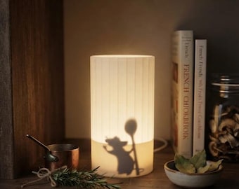 May include: A cylindrical white lamp with vertical ribbed texture, featuring a dark silhouette of a mouse holding a large spoon projected onto its surface. The lamp emits a warm glow, illuminating the surrounding space with soft light.