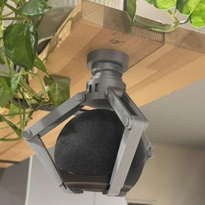 Puede incluir: Un soporte de montaje gris de estilo mecánico fijado a la parte inferior de un estante de madera, con tres brazos articulados que sujetan firmemente un altavoz inteligente esférico negro en una posición suspendida hacia abajo.