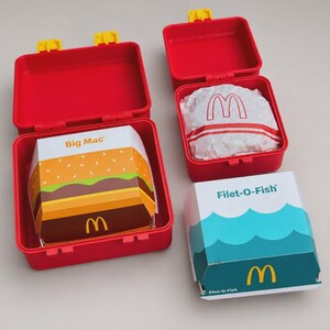 May include: Two red plastic lunch boxes with yellow latches. One box is open, containing a Big Mac burger box with a graphic of burger layers. Another red box is open, holding a wrapped sandwich with a McDonald's logo. A Filet-O-Fish box sits nearby.