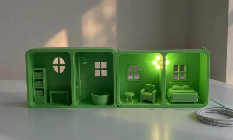 May include: A green miniature dollhouse with four rooms, each illuminated with a soft green light. The rooms include a bathroom, bedroom, and living room, with miniature furniture and fixtures. A white cord is visible.