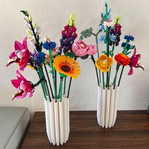 Pode incluir: Dois vasos brancos canelados contendo um arranjo colorido de flores feitas de blocos de montar de plástico. O buquê apresenta várias flores em tons de rosa, amarelo, laranja, azul, roxo e branco, apoiadas por caules verdes.