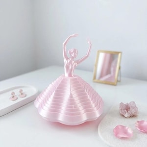 May include: A pink ballerina ornament with arms raised, wearing a layered skirt. The ornament is on a white surface, with a small mirror and decorative items in the background. The ornament is a decorative item.