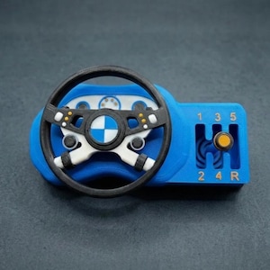 May include: A blue 3D-printed desk accessory featuring a black steering wheel with a central blue and white emblem, positioned next to a manual gear shifter gate. The gear pattern is marked with the numbers 1, 3, 5, 2, 4, and the letter R.