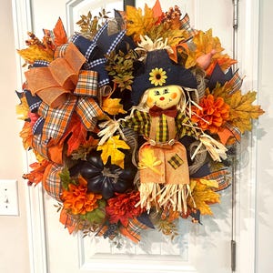 May include: A fall-themed wreath with a scarecrow figure. The wreath features orange, yellow, and brown leaves, orange and navy blue ribbons, and a black pumpkin. The scarecrow has a straw hat and a plaid shirt.