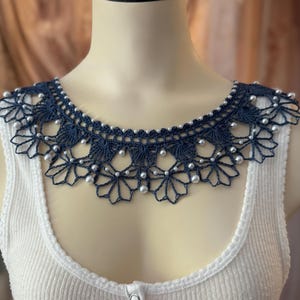 May include: A navy blue crocheted collar with a floral design and white pearl accents. The collar is displayed on a white ribbed tank top. The tank top has a scoop neck and three buttons down the front.