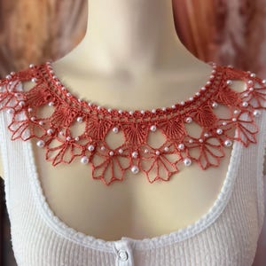 May include: A terracotta-colored, crocheted collar with a scalloped edge and adorned with small, white pearls. The collar is worn over a white ribbed tank top, creating a vintage-inspired look. The intricate lace design adds a touch of elegance.