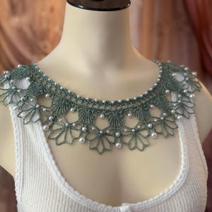May include: A sage green crocheted collar with a scalloped design and small white pearl accents. The collar is displayed on a mannequin, over a white ribbed tank top. The intricate lace-like design adds a delicate touch.