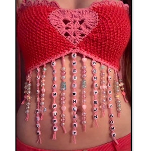 May include: A pink and red crocheted crop top with a heart-shaped design. The top has beaded fringe with letter beads spelling out the words "Sweetheart", "Cherry", "Boom", "Bun", "Honey", "Puff", "Cream", and "Plum".