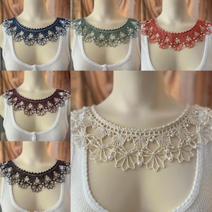 May include: A collection of handmade crochet collars in various colors, including navy blue, green, coral, burgundy, brown, and cream. Each collar features a delicate floral design and is adorned with small, white beads. The collars are displayed on mannequins.