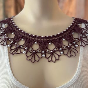 May include: A burgundy crocheted collar with a scalloped edge and white pearl accents. The collar is worn over a white ribbed shirt, creating a layered effect. The intricate crochet design adds a touch of elegance.