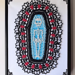 May include: A framed black and red crochet artwork featuring a skeleton in a blue coffin. The crochet piece is framed in a black frame with ornate corners.