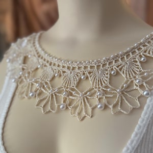 May include: Cream-colored, crocheted collar with a delicate floral and geometric design. The collar is adorned with small, round, pearl-like beads. The collar is worn over a white ribbed tank top.