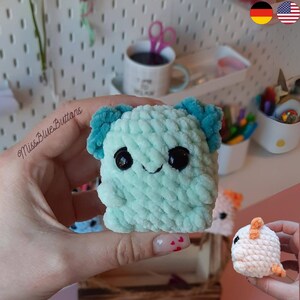 May include: A light blue crocheted plush toy with big black eyes and a small smile. The toy has teal ears and is held in a hand.