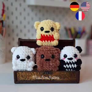 May include: A collection of handmade crochet bear toys in various colors, including yellow, white, brown, and a panda-inspired design. Each bear has black eyes and a unique expression. The bears are displayed on a decorative wooden box.