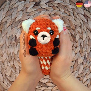 May include: A crocheted red panda plush toy with black eyes and a white nose. The toy is being held in a person's hands.