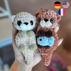 Könnte beinhalten: Zwei handgefertigte gehäkelte Otter-Plüschtiere. Eines ist hellbeige mit schwarzen Augen und einer weißen Schnauze, das andere ist braun mit blauen Augen und einem blauen Fisch. Die Plüschtiere sind weich und kuschelig, ideal für Kinder oder als einzigartiges Geschenk.