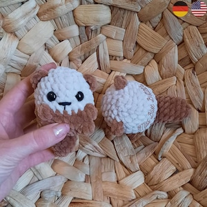 Könnte beinhalten: Zwei handgefertigte Häkeltier-Plüschotter. Einer wird in einer Hand gehalten. Die Otter sind weiß und braun mit schwarzen Augen. Ein Otter hat den Text "Miss Blue Buttons" auf dem Rücken. Die Plüschtiere befinden sich auf einem gewebten Hintergrund.