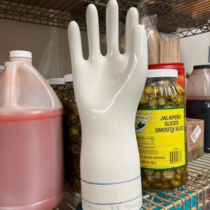 May include: A white ceramic hand-shaped display piece with a blue stripe near the base. The hand is positioned upright, with fingers extended. It is on a metal shelf, with jars of pickled jalapeños and other containers in the background.