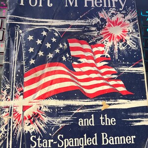 May include: A vintage book cover with the title "Fort McHenry" in white letters against a dark blue background. The cover features a waving American flag with red and white stripes and white stars, with the text "and the Star-Spangled Banner".