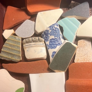 May include: A collection of various ceramic shards in shades of orange, white, blue, green, and gray. Some pieces feature patterns, including floral designs and textured surfaces. The fragments vary in size and shape, suggesting a variety of origins.