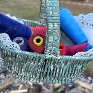May include: A woven basket painted in a distressed teal color, filled with spools of colorful thread in shades of blue, red, and pink. White lace doilies are also visible. The basket is held up, with a blurred outdoor background.