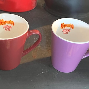 May include: Two ceramic mugs, one burgundy and one lavender, with white interiors. The mugs have the text "Kahlua ANYTHING GOES" in orange lettering inside. The mugs are on a dark surface.