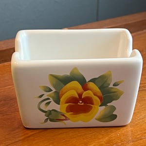 May include: A white rectangular ceramic container with a yellow and orange pansy flower design. The flower is surrounded by green leaves and buds. The container has a curved top and is sitting on a wooden surface.