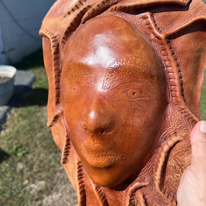 May include: A handcrafted, brown leather wall hanging featuring a sculpted face with detailed facial features. The piece has a textured surface and is surrounded by a decorative, stitched border. The artwork is held by a hand.