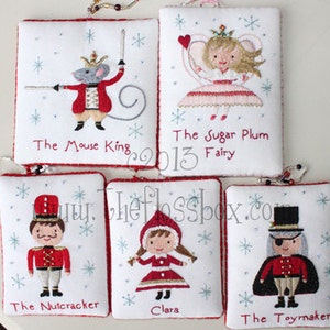 May include: Five white fabric ornaments with red trim, embroidered with characters from The Nutcracker ballet. Characters include The Mouse King, Sugar Plum Fairy, The Nutcracker, Clara, and The Toymaker. Each ornament has a beaded loop for hanging.