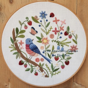 May include: Embroidered hoop art featuring a bluebird with pink breast perched on a branch with green leaves and red strawberries. The hoop also features pink, white, and blue flowers, and the text "In the Strawberry Garden The Floss Box".