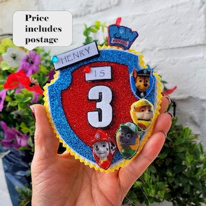 Puede incluir: Decoración de cumpleaños en forma de escudo colorido con los personajes de la Patrulla Canina. La decoración tiene un centro rojo brillante, un borde azul brillante y el número "3". El nombre "HENRY" se muestra arriba.