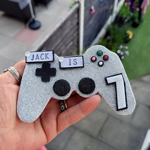 May include: A silver glittery video game controller with the text "JACK IS 7". The controller has black buttons and colourful accent buttons. A fun decoration for a gamer's birthday.