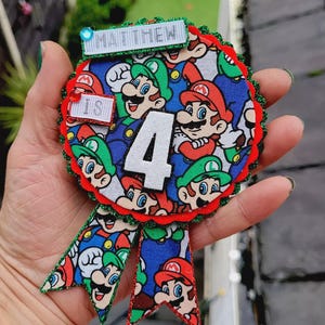 Peut inclure: Un badge rond bleu, rouge et vert avec un motif de personnages de Mario et Luigi. Le badge a un bord festonné rouge et deux rubans. Le texte "MATTHEW" et "IS 4" est affiché sur le badge.