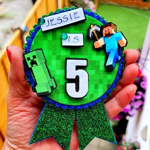 Peut inclure: Un badge d'anniversaire vert et bleu pailleté avec le nom "Jessie" et le chiffre "5". Le badge présente des personnages de Minecraft, dont un Creeper et Steve tenant une pioche. Le badge est tenu dans la main d'une personne.